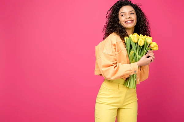 Hermosa chica americana rizada africana sonriendo mientras sostiene tulipanes amarillos aislados en carmesí - foto de stock