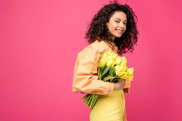 Sonriente mujer afroamericana sosteniendo tulipanes amarillos aislados en carmesí - foto de stock