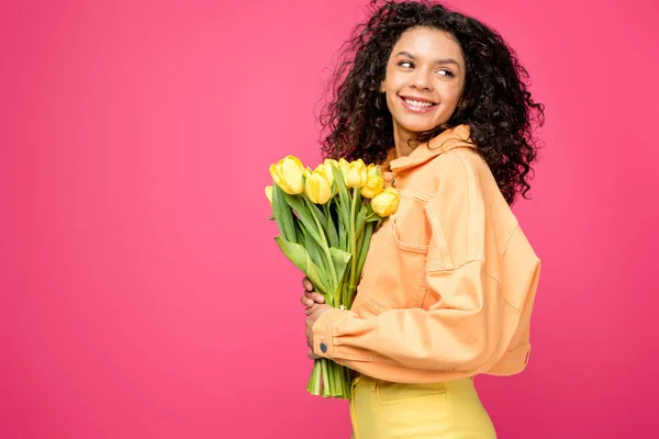 Alegre afroamericana mujer sosteniendo amarillo tulipanes aislado en carmesí - foto de stock