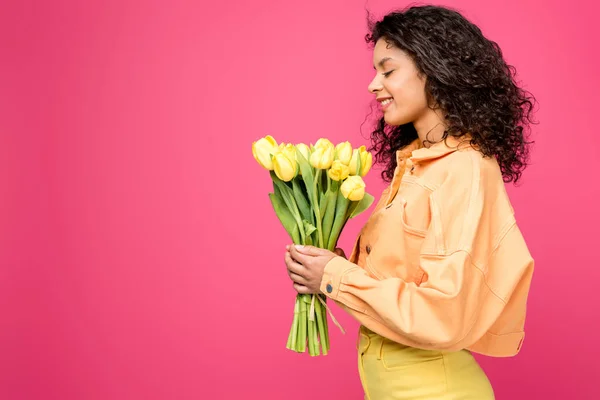 Cheerful african american woman looking at yellow tulips isolated on crimson — Stock Photo