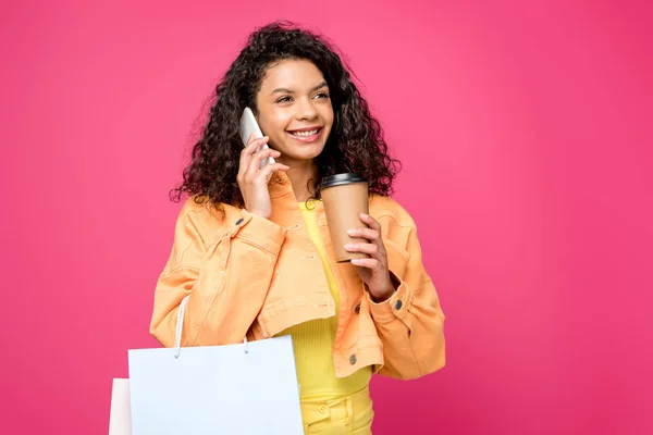 Fröhliche afrikanisch-amerikanische Frau mit Einkaufstaschen, die auf dem Smartphone spricht, während sie Pappbecher isoliert auf Purpur hält — Stockfoto