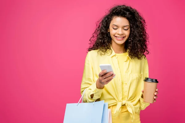 Lockige afrikanisch-amerikanische Frau mit Einkaufstaschen, die auf ihr Smartphone schaut, während sie Pappbecher isoliert auf Karminrot hält — Stockfoto