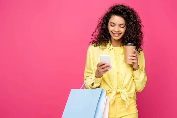 Mujer afroamericana feliz con bolsas de compras mirando el teléfono inteligente mientras sostiene la taza de papel aislada en carmesí - foto de stock