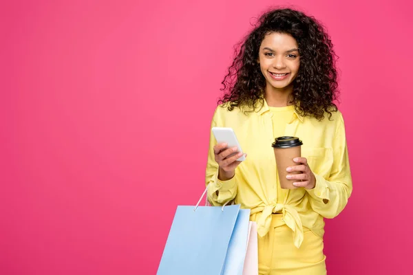 Mujer afroamericana feliz con bolsas de compras sosteniendo teléfono inteligente y taza de papel aislado en carmesí - foto de stock