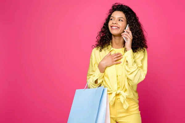 Feliz mulher afro-americana com sacos de compras falando no smartphone e de pé isolado no carmesim — Fotografia de Stock