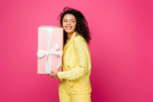 Alegre encaracolado Africano americano mulher segurando caixa de presente rosa com fita de cetim branco isolado em carmesim — Fotografia de Stock
