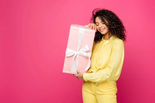 Glücklich lockige afrikanisch-amerikanische Frau hält rosa Geschenkbox mit weißem Satinband isoliert auf purpurrot — Stockfoto