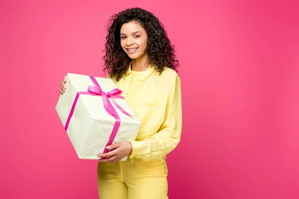 Feliz rizado africano americano mujer sosteniendo caja de regalo con cinta de satén rosa aislado en carmesí - foto de stock