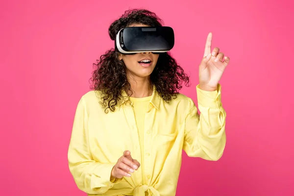 Surpreso encaracolado mulher americana africana vestindo fone de ouvido realidade virtual e apontando com os dedos isolados no carmesim — Fotografia de Stock