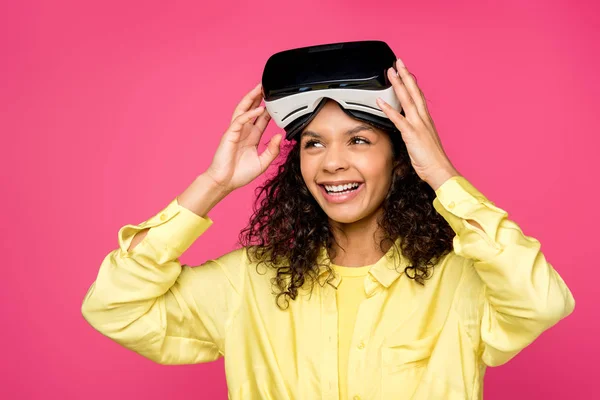 Feliz rizado mujer afroamericana con auriculares de realidad virtual y sonriendo aislado en carmesí - foto de stock