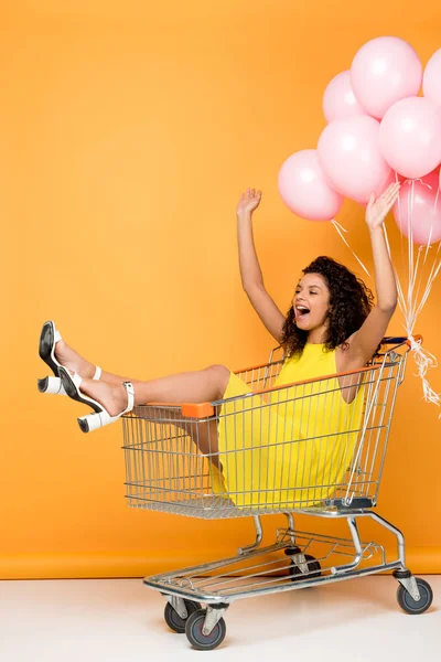 Cheerful african american woman screaming while sitting in trolley near pink air balloons on orange — Stock Photo