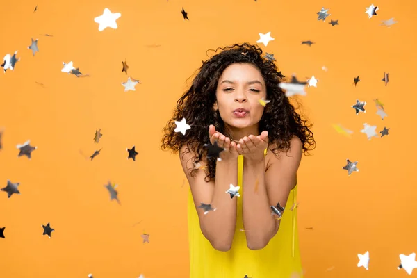 Happy curly african american girl in yellow dress sending kiss near shiny confetti stars on orange — Stock Photo