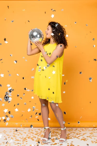 Atraente encaracolado menina americana africana segurando bola de discoteca perto de estrelas de confete brilhantes em laranja — Fotografia de Stock