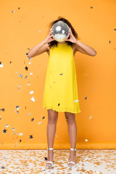 Chica afroamericana en vestido amarillo cubriendo la cara con bola disco cerca de estrellas de confeti brillantes en naranja - foto de stock