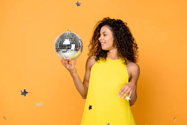 Feliz rizado africano americano mujer mirando disco bola cerca brillante confeti estrellas en naranja - foto de stock