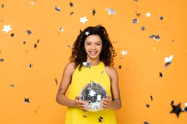 Selective focus of attractive curly african american girl holding disco ball near shiny confetti stars on orange — Stock Photo