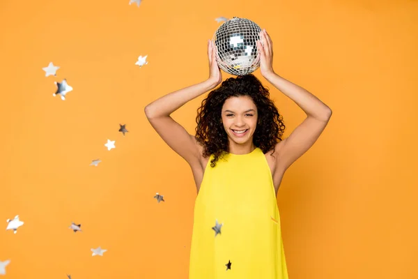 Enfoque selectivo atractivo rizado chica afroamericana sosteniendo bola disco por encima de la cabeza cerca de estrellas de confeti brillantes en naranja - foto de stock