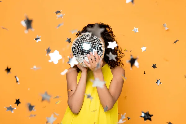 Selective focus of african american girl in yellow dress covering face with disco ball near shiny confetti stars on orange — Stock Photo