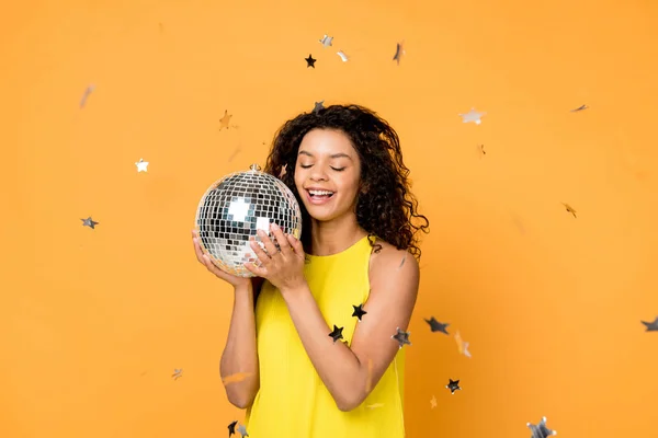 Rizado africano americano mujer celebración disco bola cerca brillante confeti estrellas en naranja - foto de stock