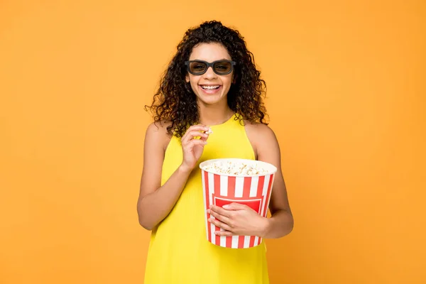 Joyeuse femme afro-américaine bouclée tenant seau de maïs soufflé et souriant dans des lunettes de soleil isolées sur orange — Photo de stock