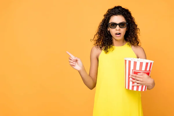 Surprised curly african american woman holding popcorn bucket and pointing with finger isolated on orange — Stock Photo