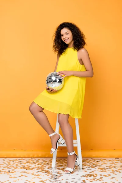 Allegra donna riccia afroamericana che tiene la palla da discoteca mentre siede sulla sedia su arancione — Foto stock