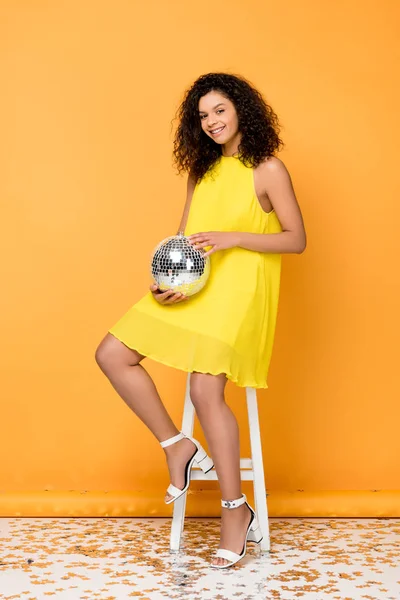 Joyeuse femme afro-américaine bouclée tenant boule disco tout en étant assis sur une chaise sur orange — Photo de stock