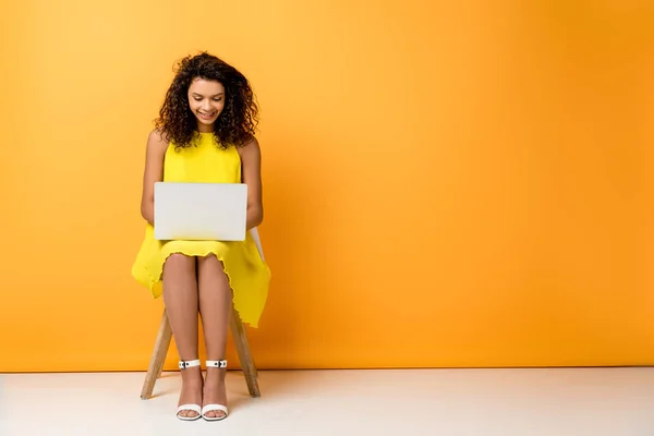 Heureux frisé afro-américaine femme assise en robe jaune sur chaise et en utilisant un ordinateur portable sur orange — Photo de stock