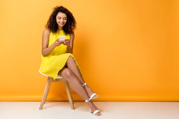 Happy curly african american woman sitting in yellow dress on chair and using smartphone on orange — Stock Photo