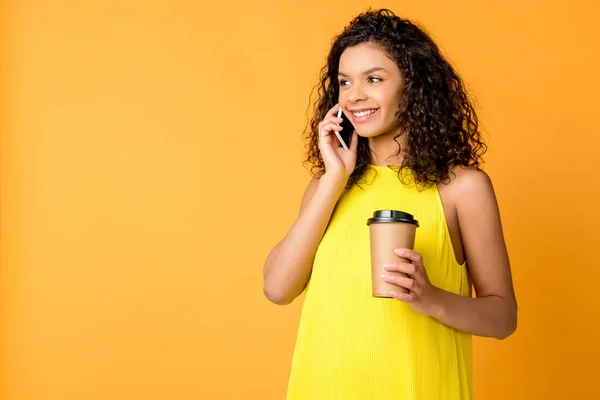 Felice donna afro-americana riccia che parla su smartphone mentre tiene isolata la tazza di carta su arancione — Foto stock