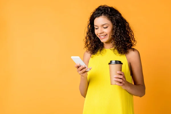 Feliz rizado mujer afroamericana usando teléfono inteligente mientras sostiene taza de papel aislado en naranja - foto de stock
