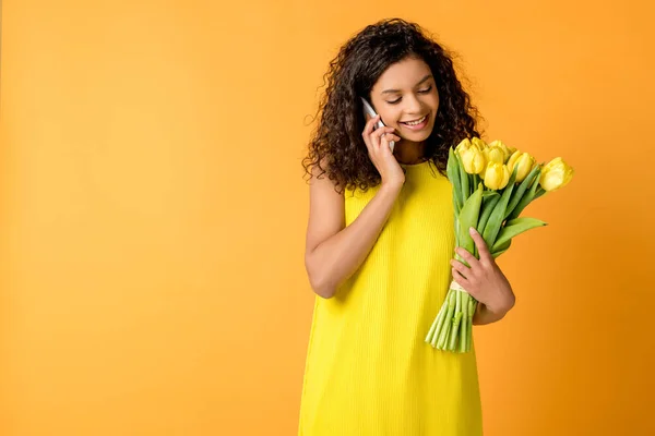 Glücklich lockige afrikanisch-amerikanische Mädchen spricht auf dem Smartphone, während sie gelbe Tulpen isoliert auf orange halten — Stockfoto