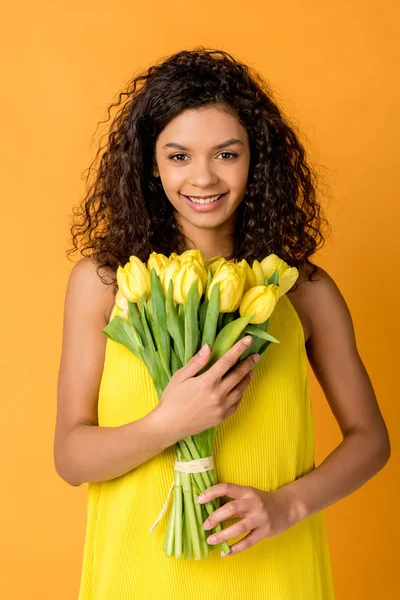 Glücklich lockige afrikanisch-amerikanische Frau hält gelbe Tulpen isoliert auf orange — Stockfoto