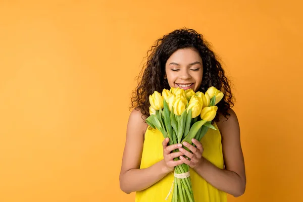 Glücklich lockige afrikanisch-amerikanische Frau riecht gelbe Tulpen isoliert auf orange — Stockfoto