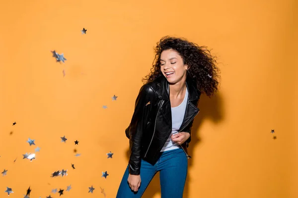 Happy african american girl standing near shiny confetti stars on orange — Stock Photo