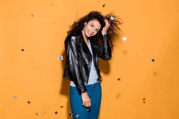 Smiling african american girl standing near shiny confetti stars on orange — Stock Photo