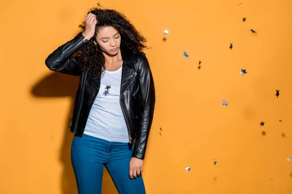 Curly african american girl standing near shiny confetti stars on orange — Stock Photo