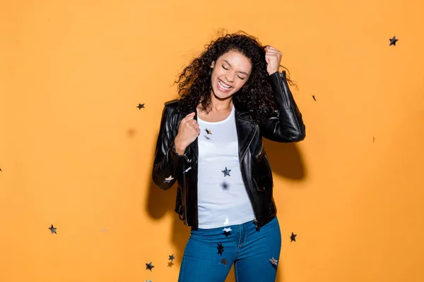 Excited pretty african american girl standing near shiny confetti stars on orange — Stock Photo