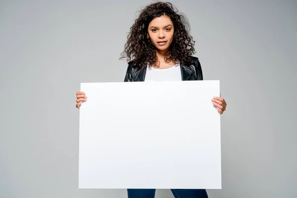 Linda menina americana africana encaracolado segurando cartaz em branco, enquanto de pé isolado em cinza — Fotografia de Stock