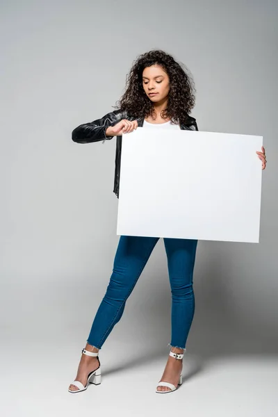 Hermosa chica afroamericana rizada sosteniendo pancarta en blanco mientras está de pie en gris - foto de stock