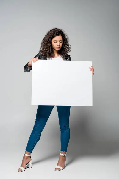 Chica afroamericana bastante rizada sosteniendo pancarta en blanco mientras está de pie en gris - foto de stock