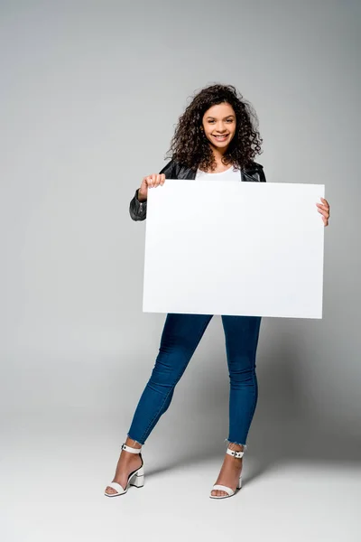 Alegre rizado africano americano chica sosteniendo en blanco cartel mientras de pie en gris - foto de stock