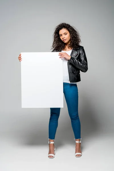 Красива кучерява афроамериканська жінка тримає порожній плакат, стоячи на сірому — стокове фото