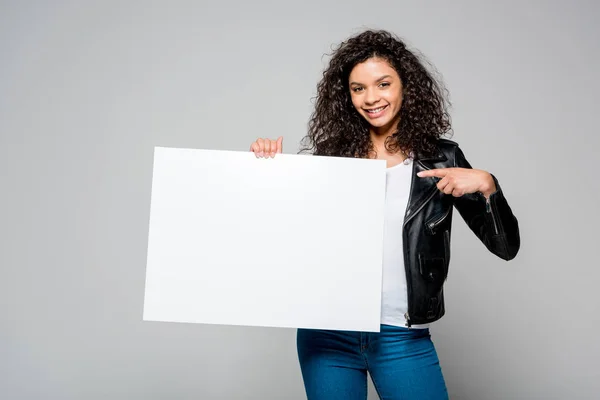 Счастливая африканская американская молодая женщина, указывающая пальцем на пустой плакат, стоящая изолированная на сером — стоковое фото