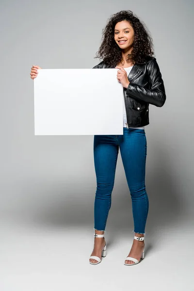 Feliz afroamericana joven mujer sosteniendo cartel en blanco en gris - foto de stock