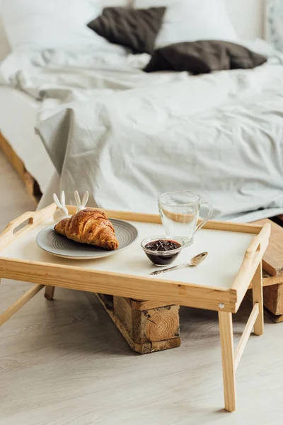 Селективный фокус деревянного подноса с круассаном, вареньем и чашкой в спальне с копировальным пространством — стоковое фото