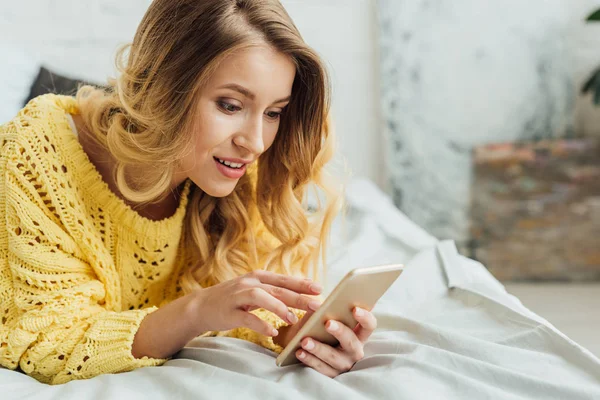 Schönes Mädchen im Strickpullover auf dem Bett liegend und Smartphone benutzend — Stockfoto