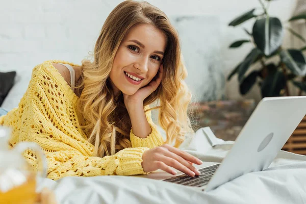 Foco seletivo de bela menina sorridente deitada na cama e olhando para a câmera enquanto usa laptop em casa — Fotografia de Stock