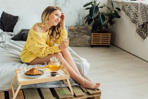Bella ragazza seduta vicino al vassoio con cibo e tè versante in tazza durante la colazione in camera da letto — Foto stock