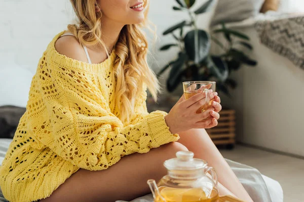 Abgeschnittene Ansicht einer jungen Frau im Strickpullover, die auf dem Bett sitzt und eine Tasse Tee in der Hand hält — Stockfoto